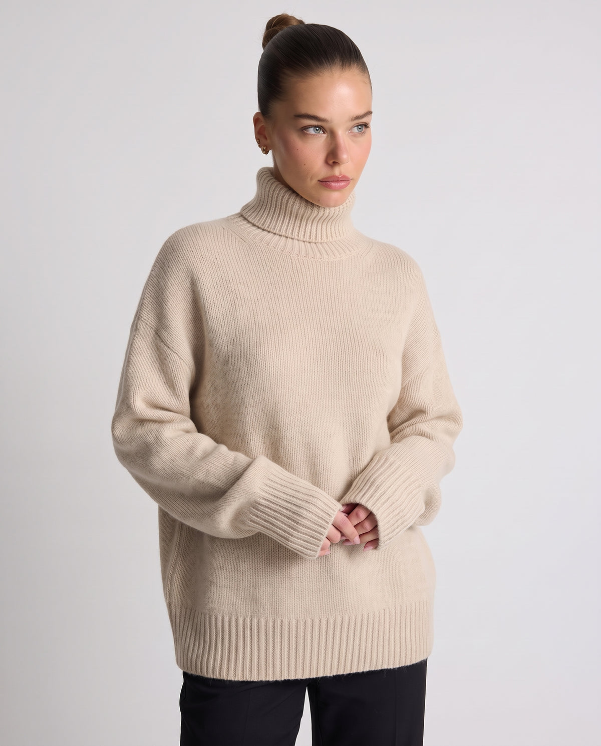 A woman wearing a beige ribbed turtleneck sweater with oversized sleeves, paired with black pants, standing against a light gray studio background.